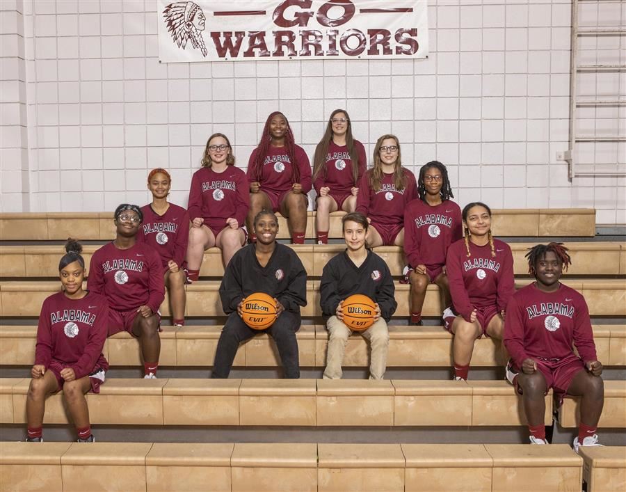 2022-23 ASD girls varsity basketball team sit in an upsidedown v shape in the gym bleachers.